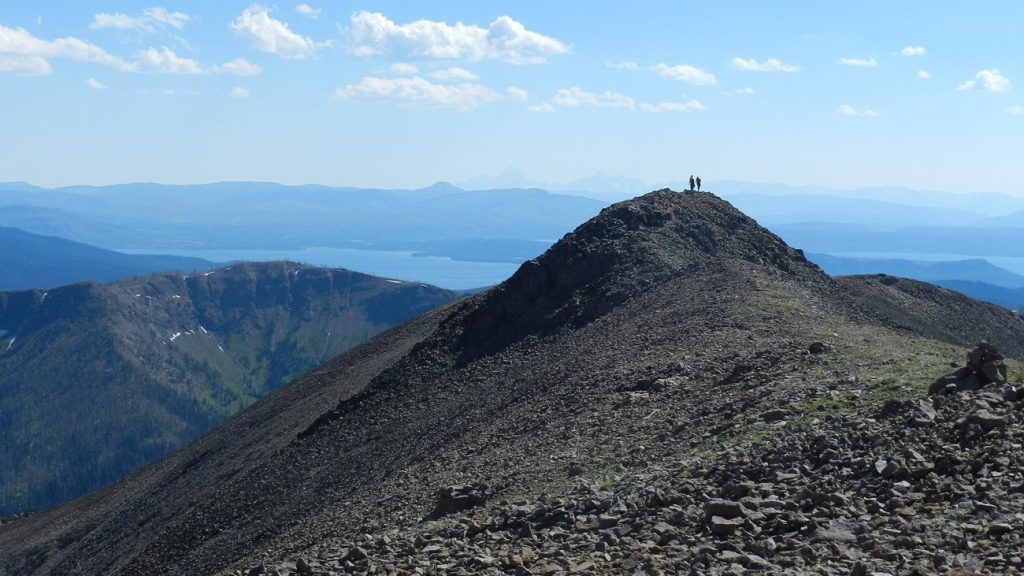 Two hikers stand on the rocky summit of Avalanche Peak with mountain ranges in the distance.