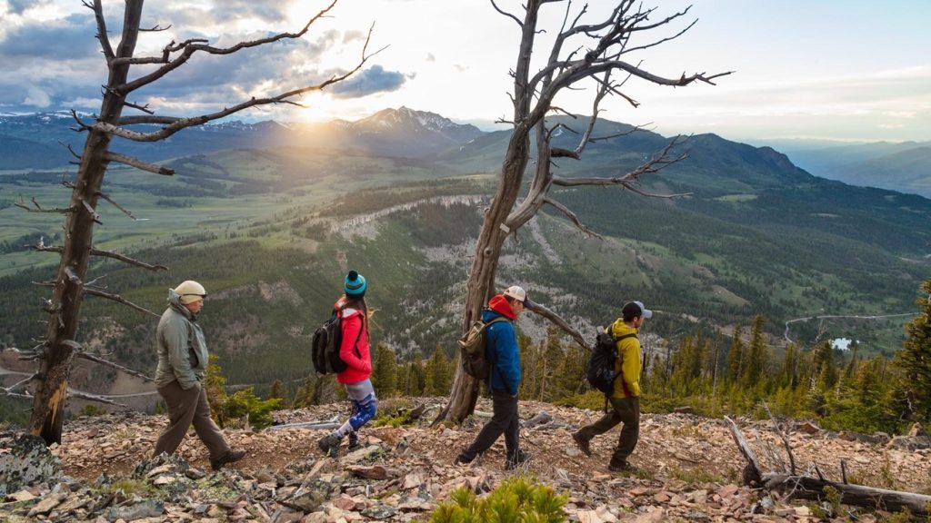 Four hikers cross a rocky Bunsen Peak slope at sunset with Yellowstone mountains and valleys behind them.