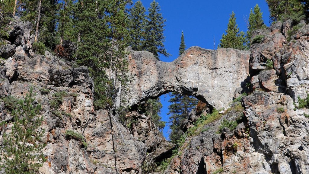 Yellowstone's Natural Bridge spans a rocky opening in a forested rhyolite cliff beneath a deep blue sky.