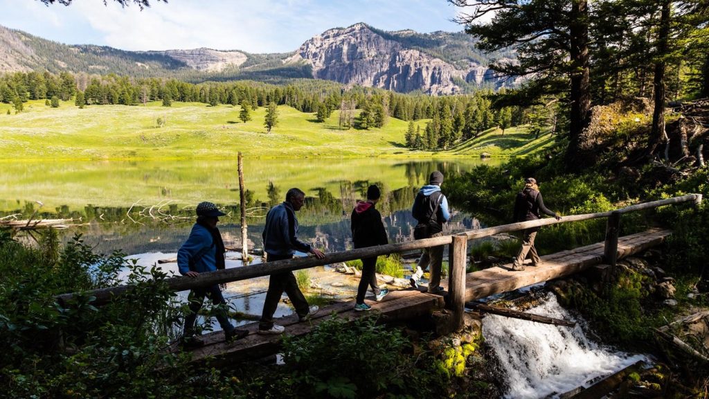 Hikers cross a small wooden bridge beside Trout Lake with green meadows and rocky hills beyond.